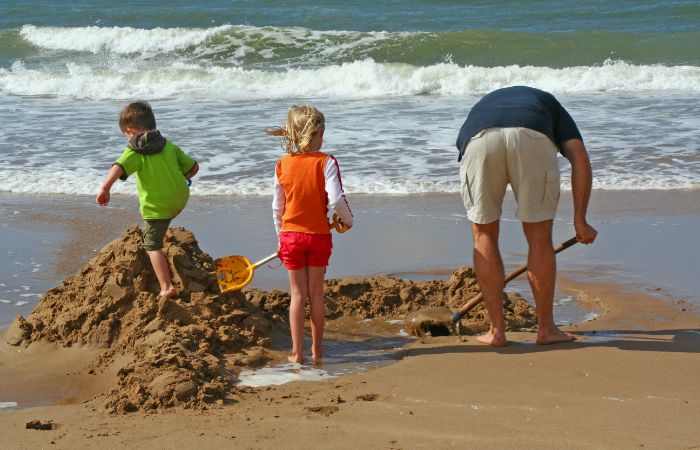 niño, niña y padre construyen un muro de arena frente a las olas del mar Juegos en la playa: muro contra las olas