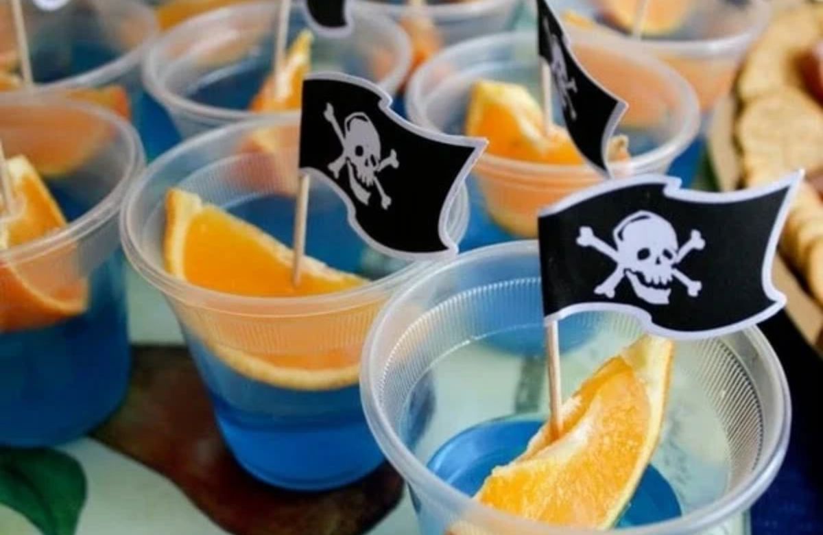 Barquitos piratas hechos con gajos de naranja flotando en vasos con un mar azul Fiesta infantil de piratas: vasos de bebida decorados